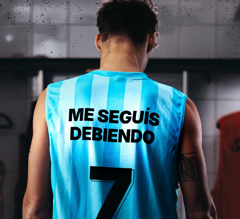 De espaldas en un vestuario, un pibe con camiseta celeste número 7 que en vez del nombre dice ME SEGUÍS DEBIENDO.