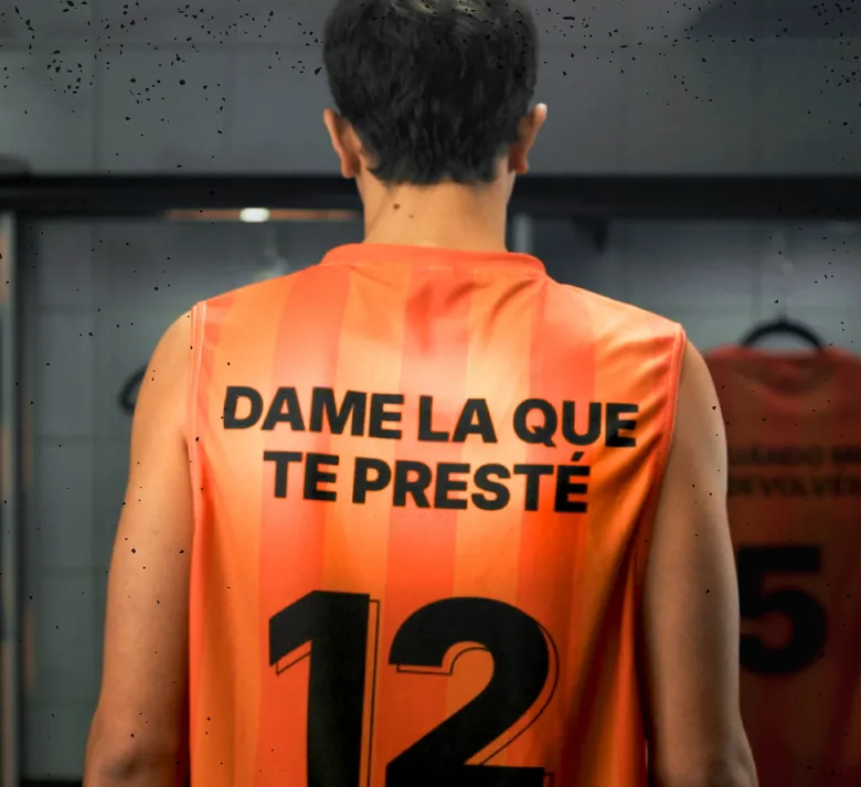 De espaldas, un pibe con camiseta naranja número 12 que dice DAME LA QUE TE PRESTÉ.